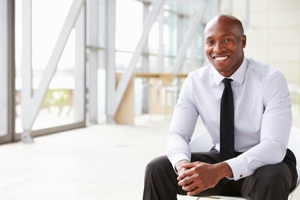 A smiling man in a white shirt and black tie seated in a bright office