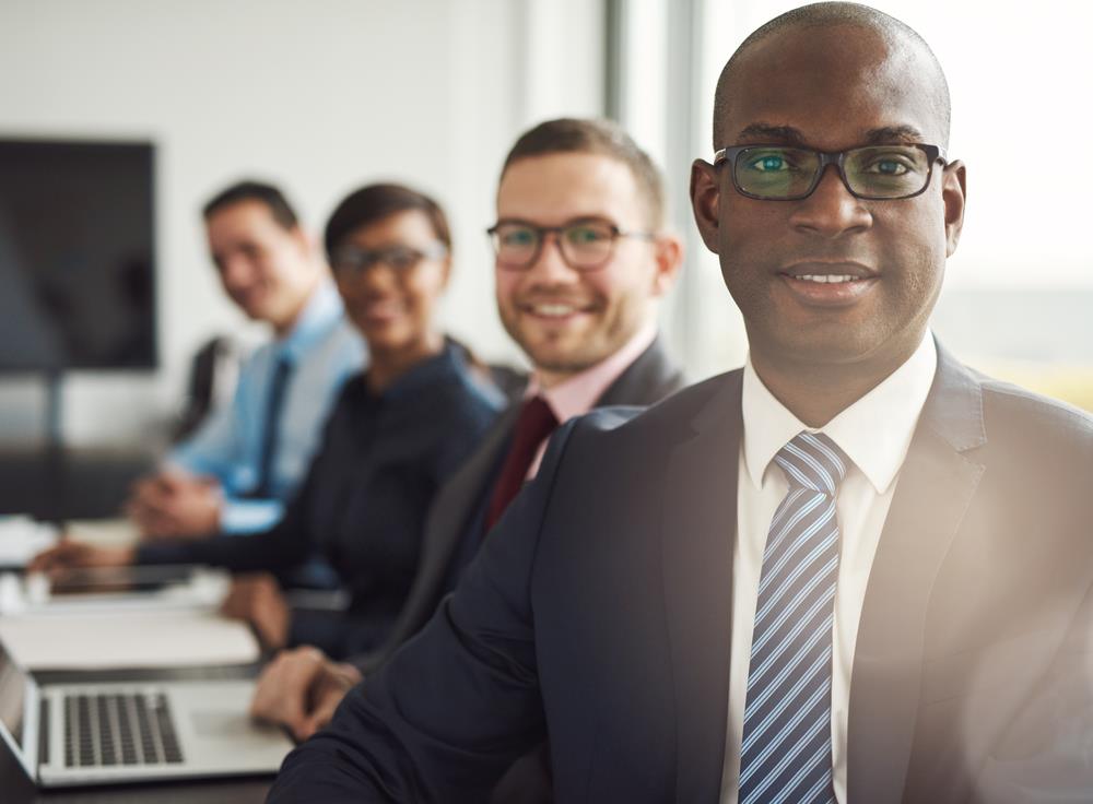 A smiling man in a suit and tie in front of colleagues seated at a boardroom table