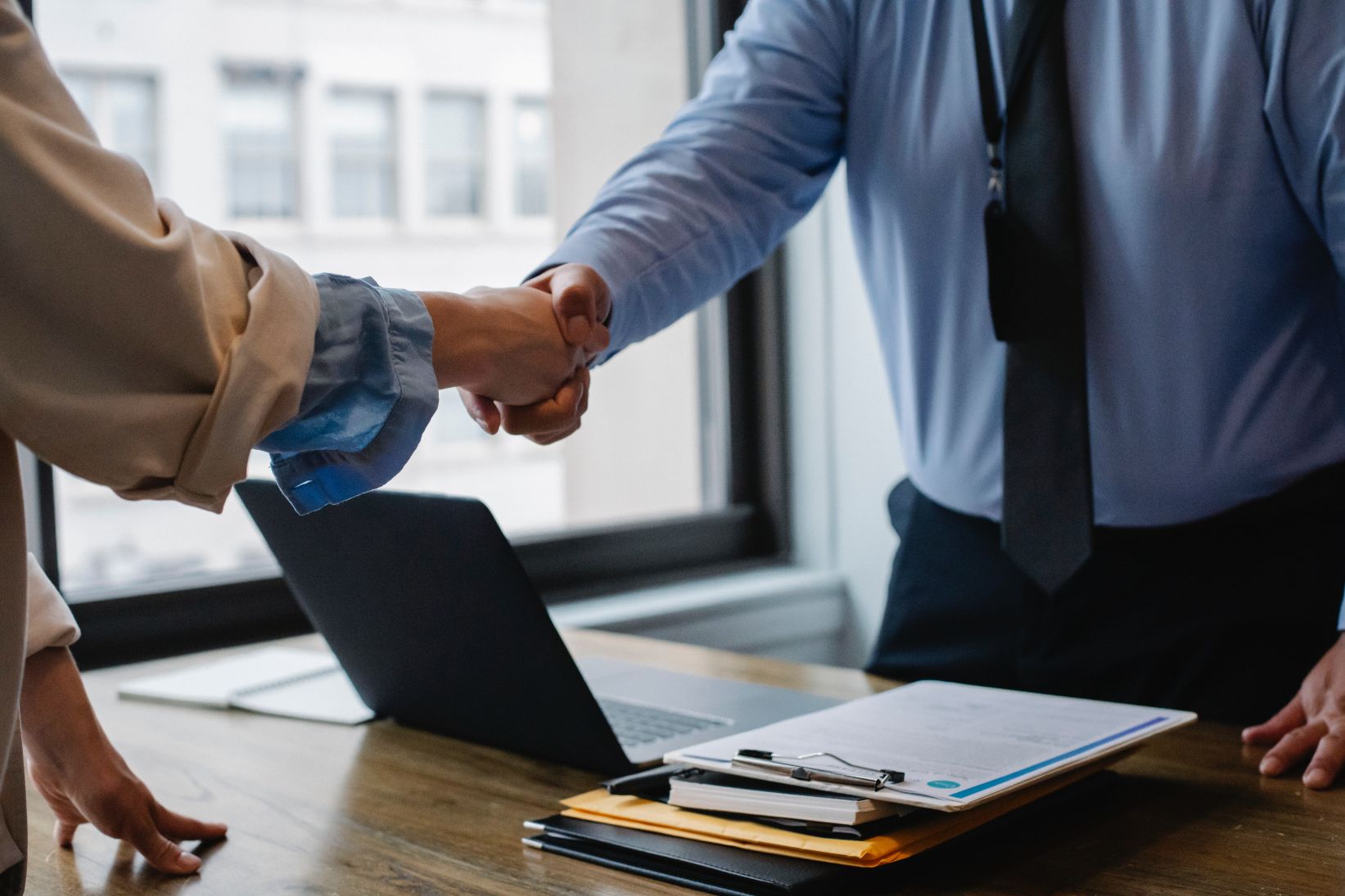 Two people shaking hands across a desk with a laptop and documents