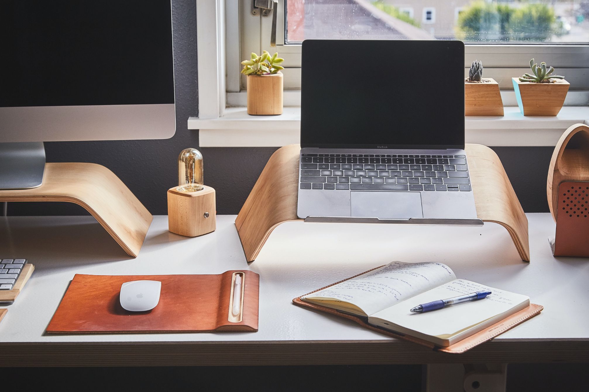 A tidy home desk with a laptop, notebook and potted plants by a window
