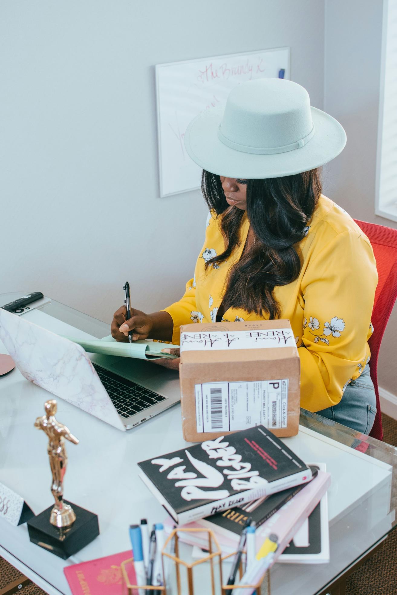 A woman in a hat and yellow jacket working at a desk with parcels and a laptop