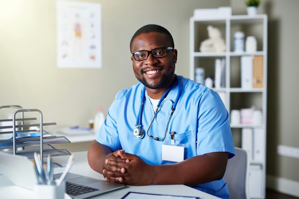 A smiling male nurse in scrubs seated at a clinic desk