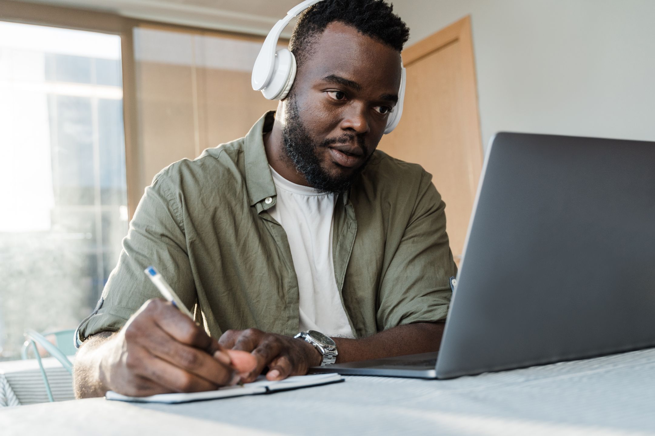 A man with headphones writing notes beside a laptop