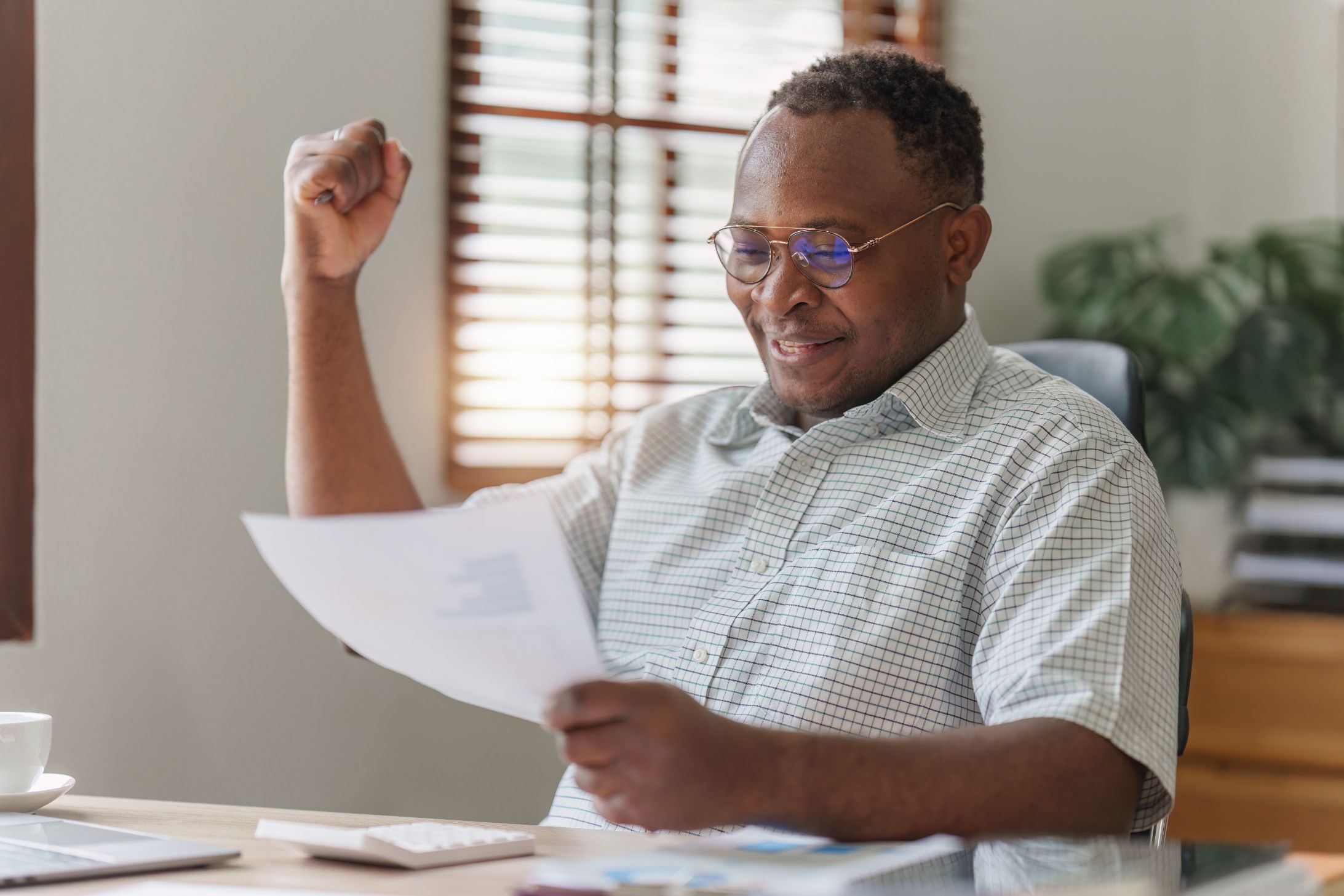 A man in glasses raising a fist in celebration while reading a letter at his desk