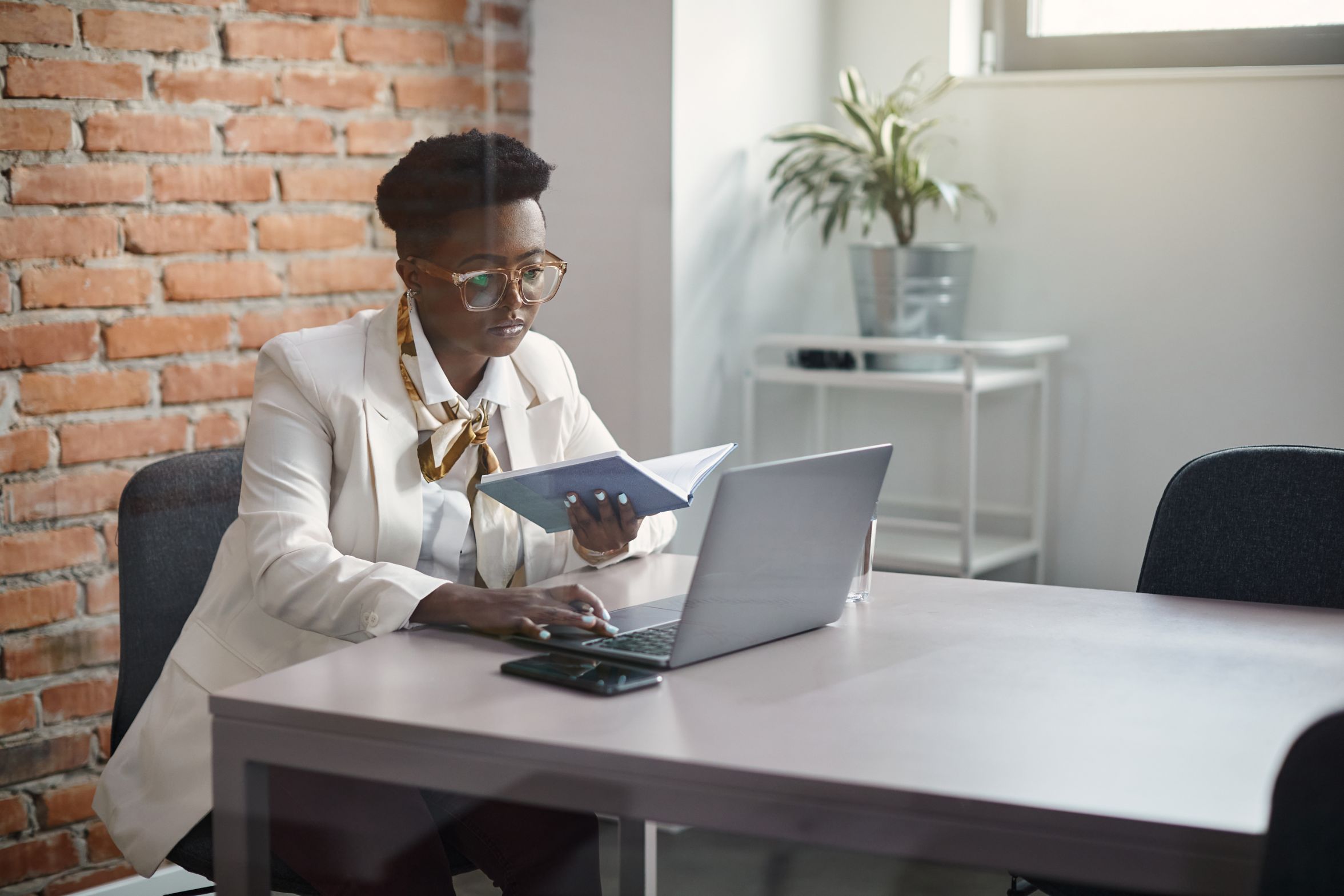 A woman in a white blazer reading a tablet at a desk with a laptop