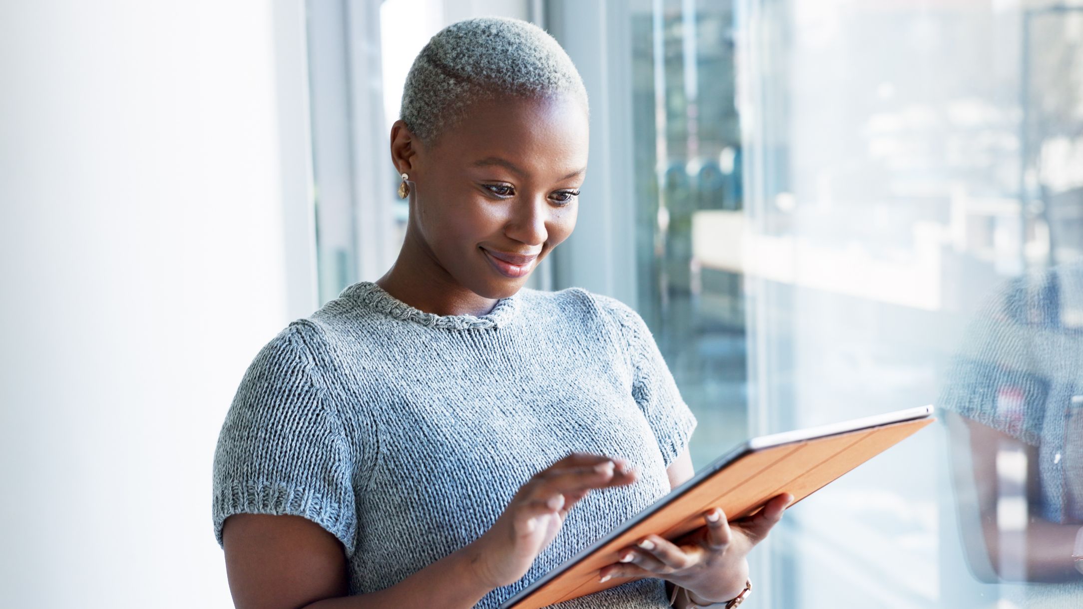 A woman with short hair smiling at a tablet by a window