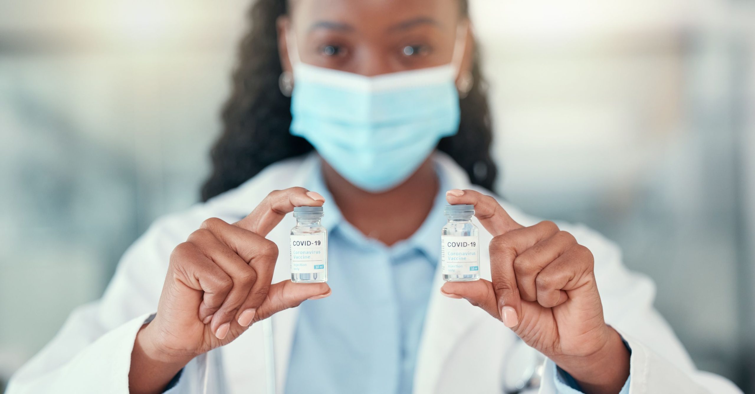 A health worker in a mask holding up two vaccine vials