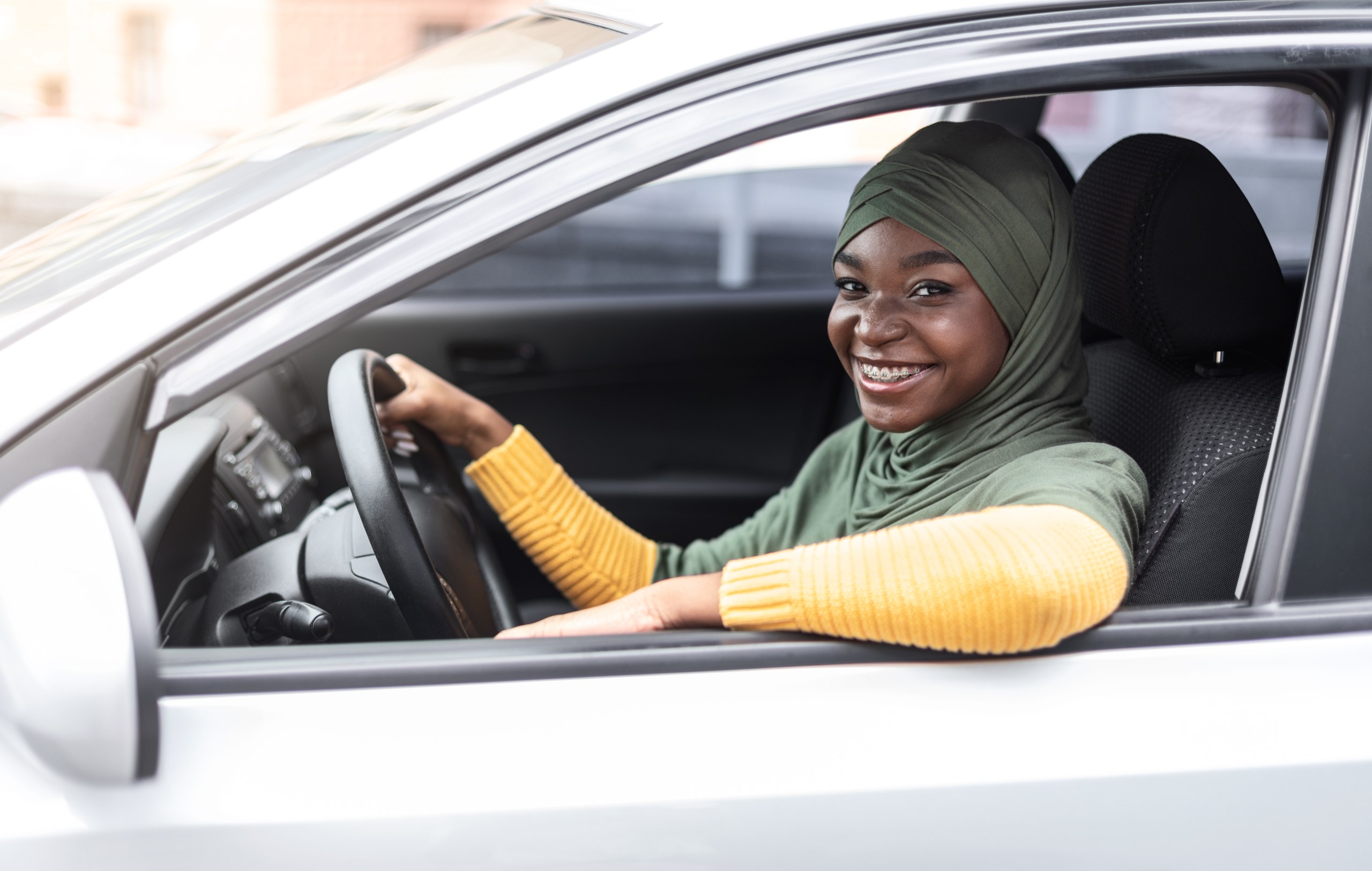 A smiling woman in a headscarf at the wheel of a white car