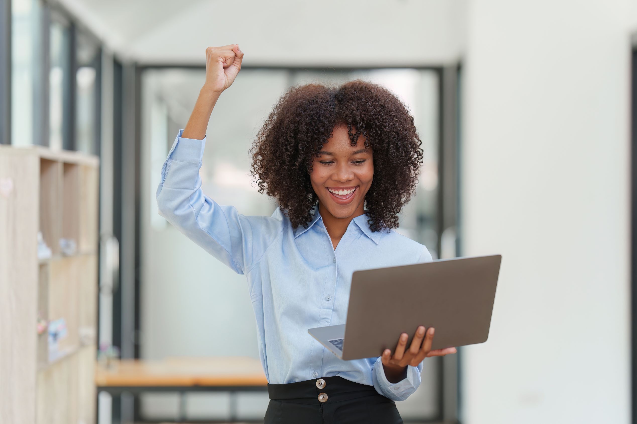 A smiling woman raising a fist in celebration while holding a laptop