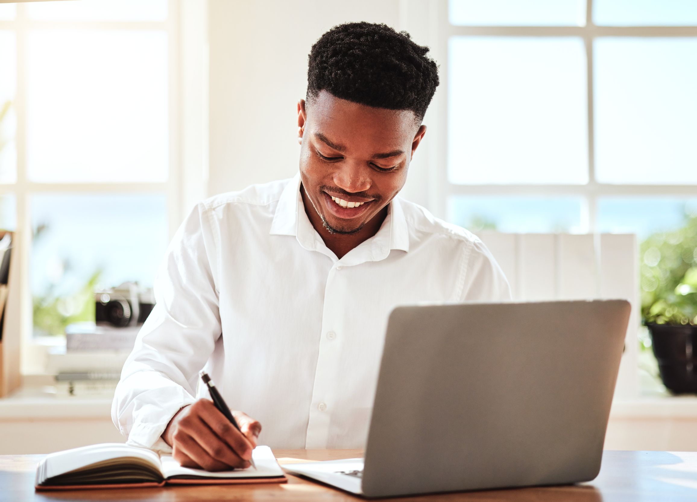 A smiling young man writing in a notebook beside a laptop