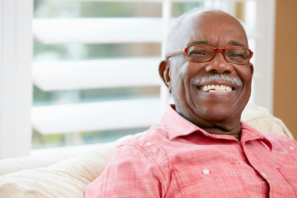 A smiling older man with glasses and a moustache seated on a sofa