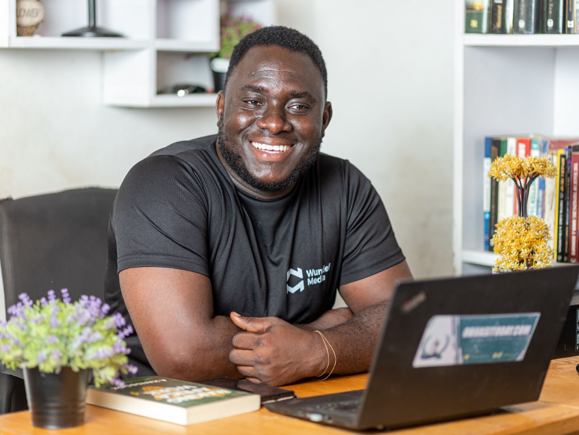 A smiling man at a home desk with a laptop and bookshelves