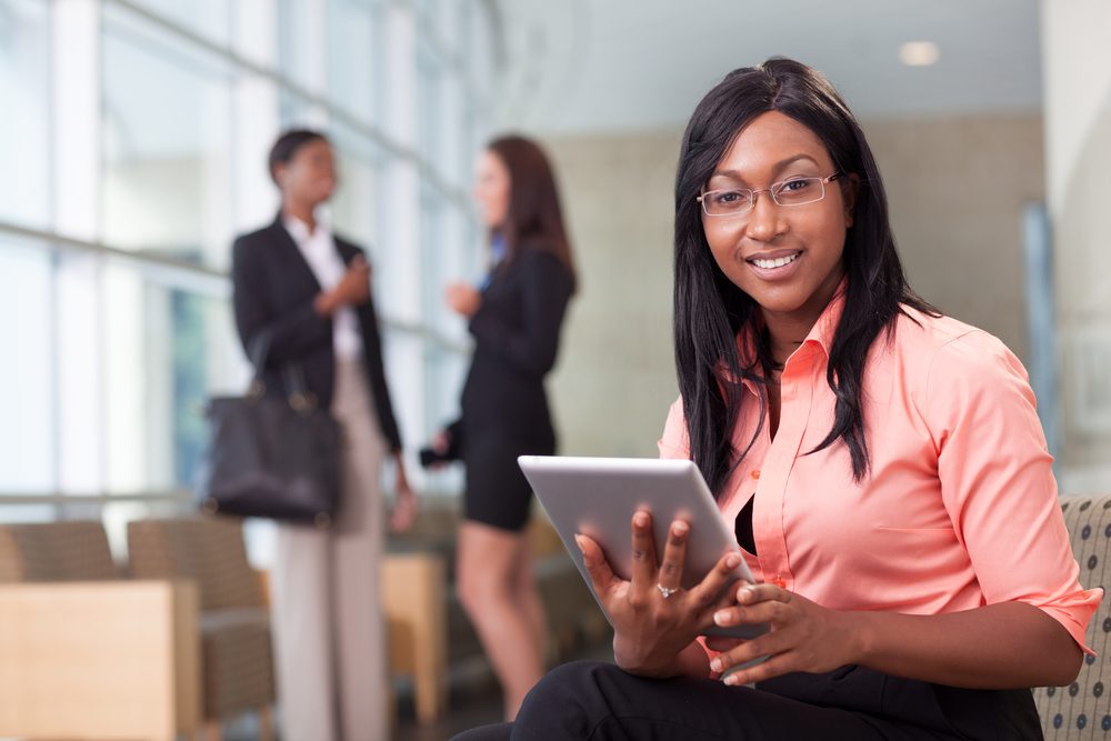 A smiling woman with a tablet in an office lobby, colleagues behind her