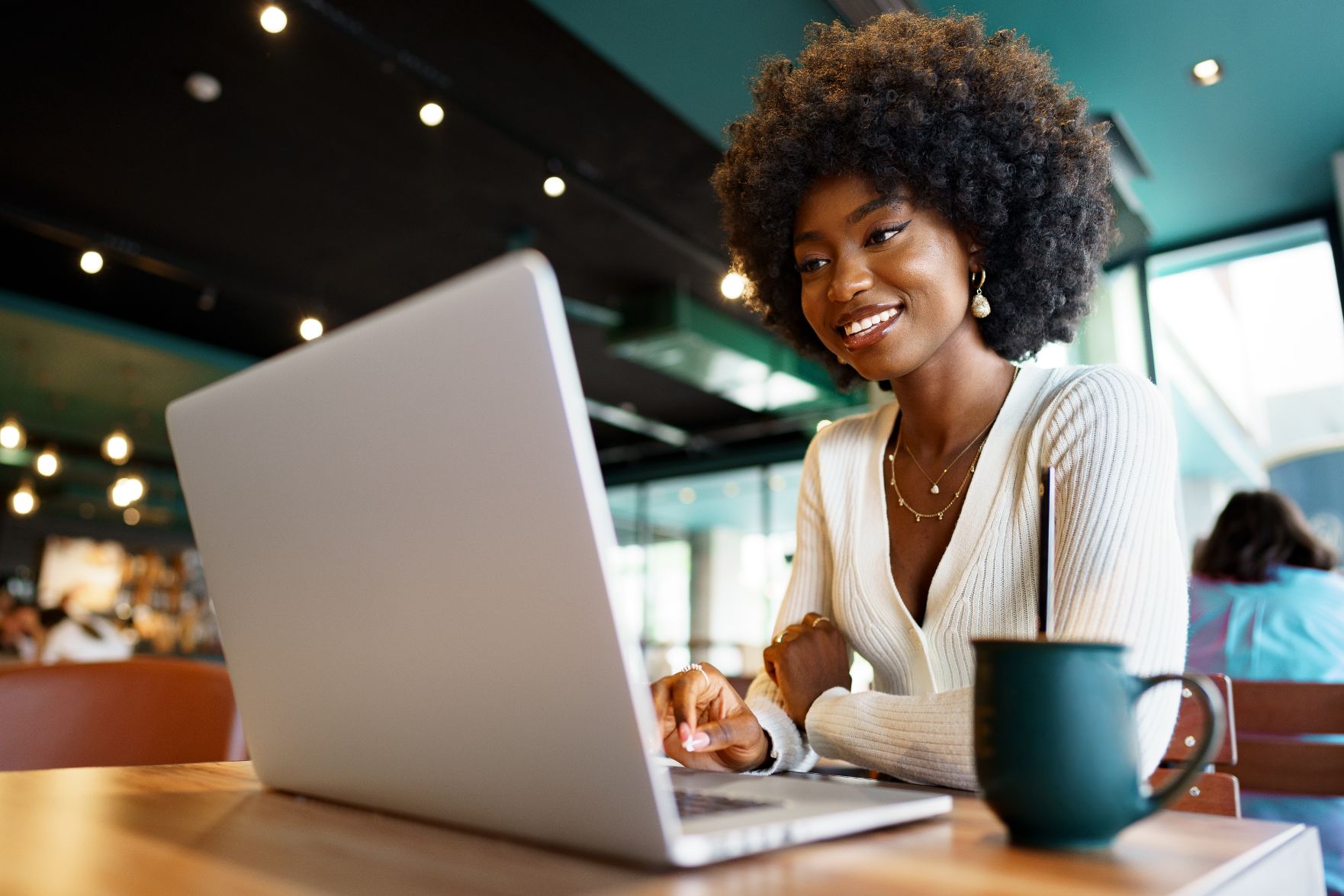 A smiling young woman with a laptop and a mug at a café table