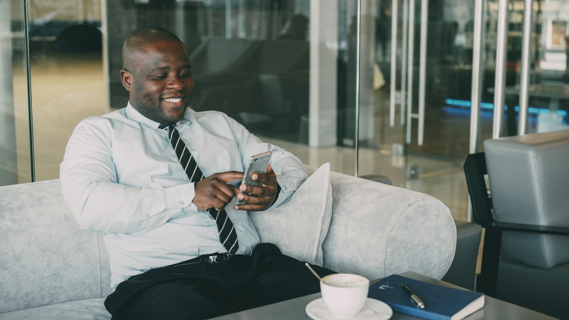 A man in a shirt and tie smiling at his phone on an office sofa
