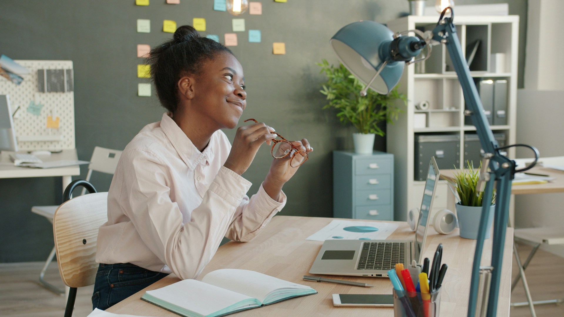 A smiling woman at a desk in a bright home office with a laptop and notebook