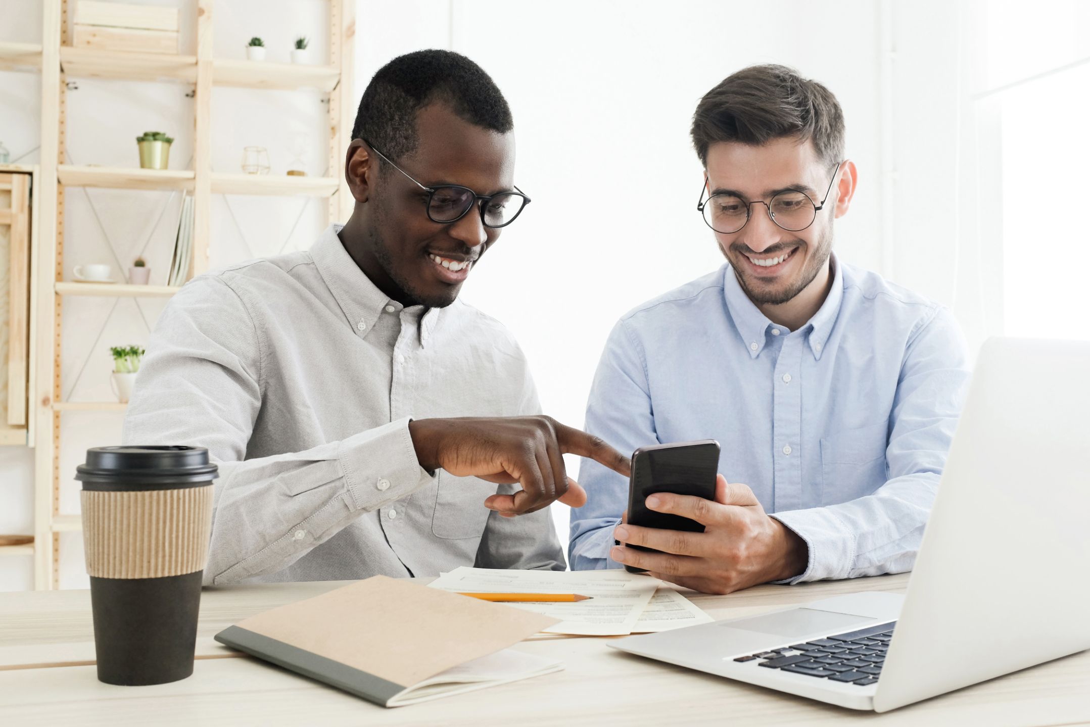 Two colleagues smiling over a phone beside a laptop and coffee